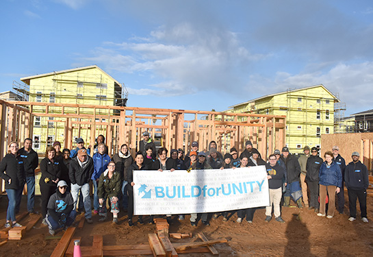 A group of about 40 people, most wearing coats and many wearing hats, stand together at a homebuilding site, behind a large white banner