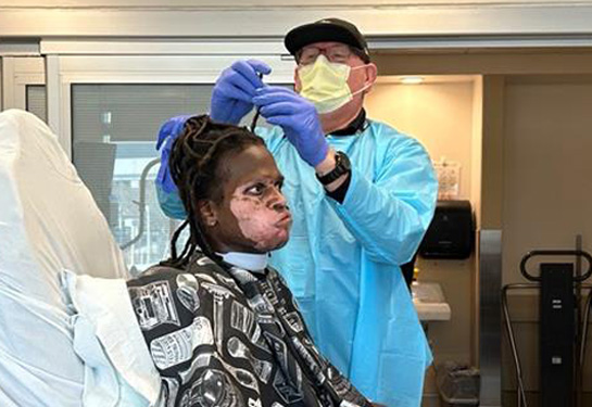Man wearing mask and hospital gown stands next to and repairs a dreadlock on a recovering burn patient sitting in a chair.