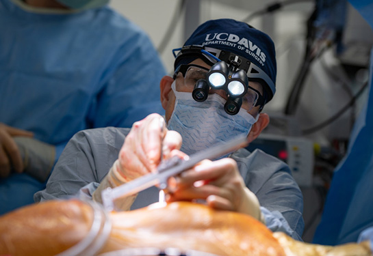 Surgeon operating wearing mask, blue UC Davis head cover and glasses with a light