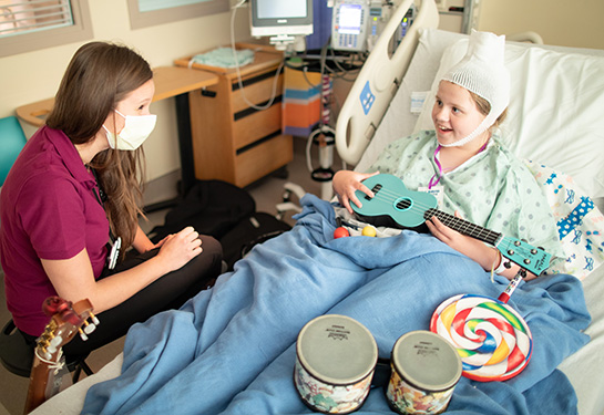 Woman sits by the bedside of a girl with a bandage on her head playing a ukulele in her hospital bed.