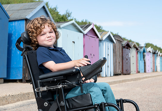 A six year old boy sitting in wheelchair in front of colorful painted beach huts, looking at camera, smiling