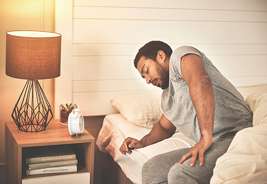 A man wearing gray sweatpants and a gray t-shirt sits with feet on the floor next to the bed and eyes closed. His nightstand holds an alarm clock