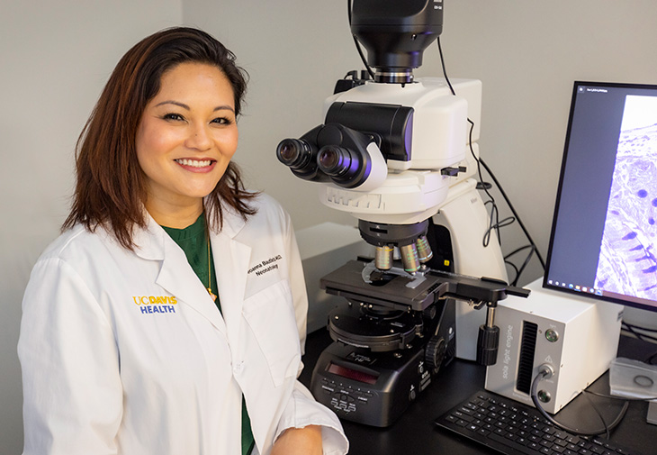 A smiling woman wearing a lab coat, sitting in front of an electronic microscope, a computer, and a monitor