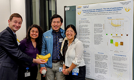 A man resembling Brad Pitt offers bananas to three brilliant medical students demonstrating their MAD Skills in front of their colorful poster