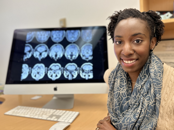 A smiling young woman with short brown hair in a tan sweater and a blue scarf is in front of a computer monitor showing a series of MRI brain scans. 