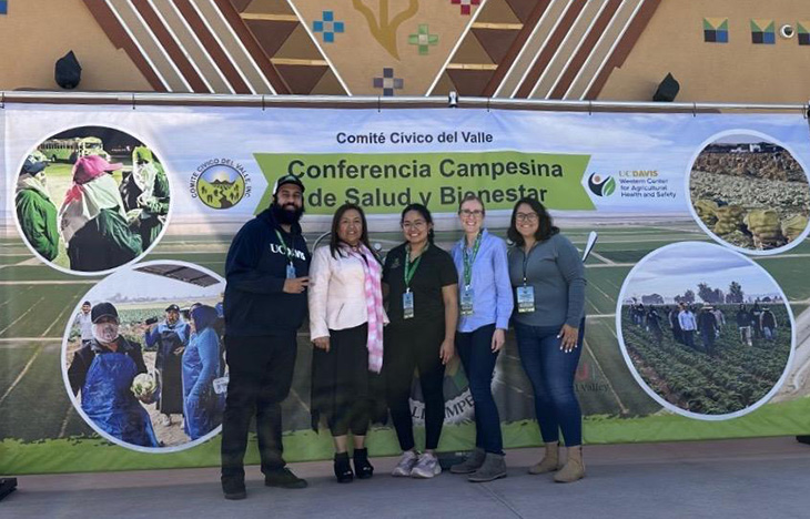 Five people stand in front of a large sign that says Conferencia Campesina de Salud y Bienestar.