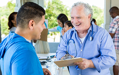 Man with stethoscope over his shoulders speaks to a patient at an outdoor event.