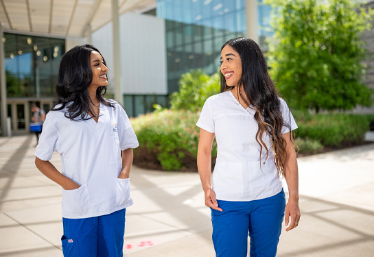 two nursing students wearing scrubs stand outside building smiling at each other