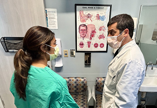 A female high school student in scrubs and facemask, left, listens to a male doctor in white scrubs and facemask