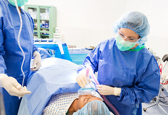 A young patient is being sedated while laying on operating table. A female anesthesiologist is using medical equipment to sedate patient.