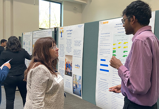 A room with posters on walls and presenters explaining to their work to attendees. To the right, a student in eyeglasses stands by his poster 
