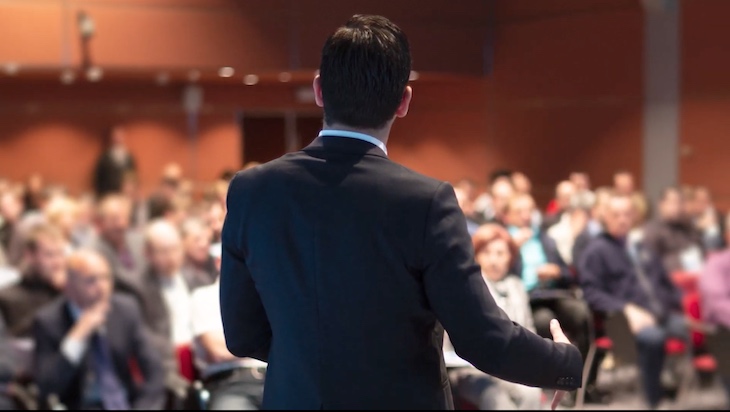 A man in a suit is seen from behind talking to a large crowd