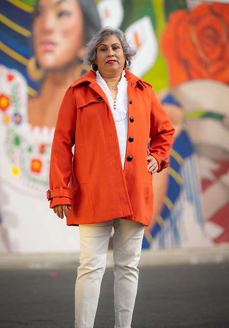 Grey-haired Latina wearing an orange coat and white shirt and pants in front of mural.