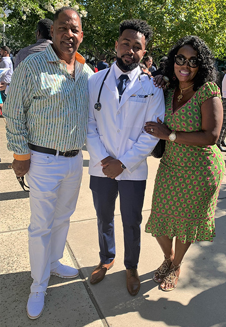Medical student Brandon Coleman in white coat is flanked by his father and mother, standing in the courtyard at UC Davis Health 