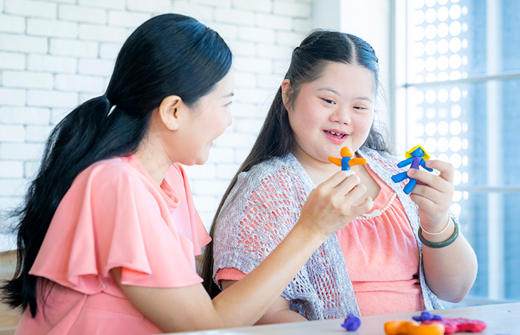 A mother with long black hair and daughter with long black hair, who has down syndrome, sit at a table playing with colorful plastic toys. 