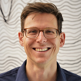 Man with light brown hair and glasses and navy button up shirt in front of abstract white and black lined background.