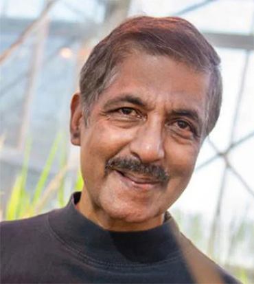 a smiling middle-aged man with a moustache and wearing a black mock-turtleneck shirt stands in a greenhouse. 