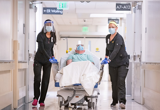 Two female nurses wearing black scrubs push patient in bed down the hallway