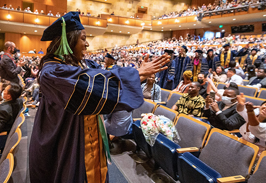 A female medical student in blue gown and cap gestures to supporters in the audience of Mondavi Center during Commencement 2022