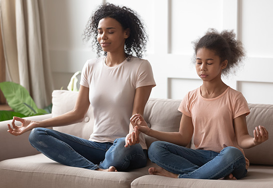 African American mother and her young daughter sit on a couch, eyes closed, legs crossed, arms out at side and palms raised, to do breathing exercise