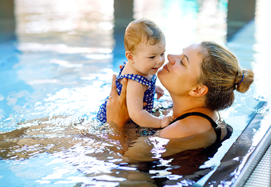 Blonde woman with hair in a bun holds toddler girl in her arms with short light brown hair in indoor swimming pool 