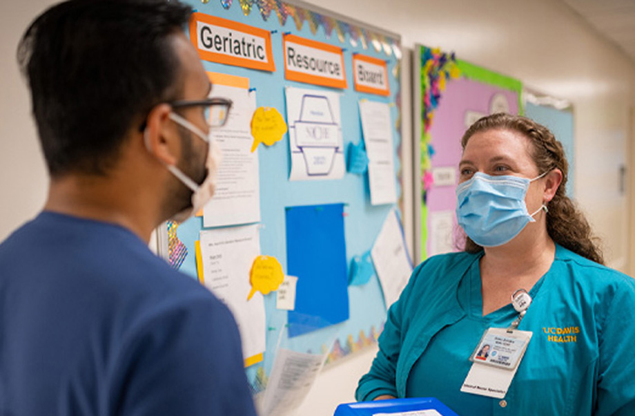  Nurse wearing scrubs and mask stands in front of bulletin board and talks to nurse colleague who looks at her and whose back is facing the camera