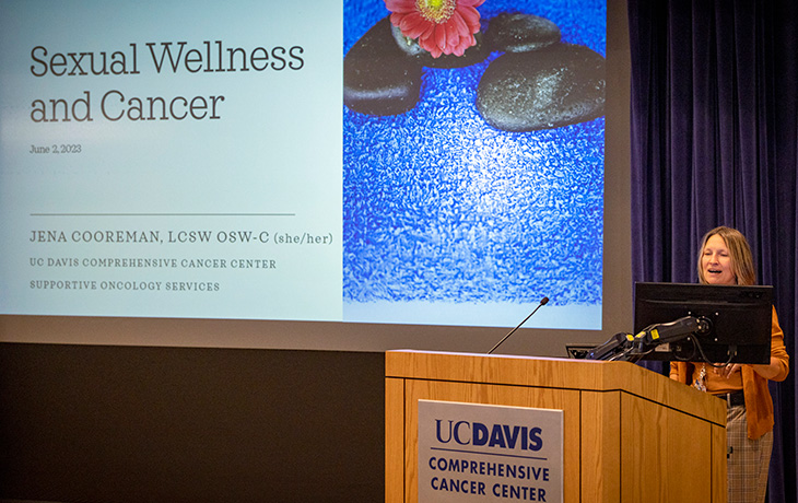 Woman at podium inside auditorium with large screen behind her that says “Sexual Wellness after Cancer”