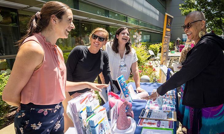 African American woman talking with three women behind a table with pamphlets and other material on a table that says UC Davis Comprehensive Cancer Center