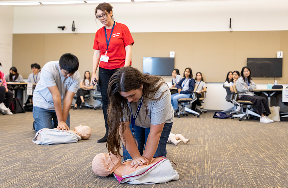 A woman in red polo shirt stands and gestures as two teenaged students, a boy and a girl, practice CPR on manikins on the floor 