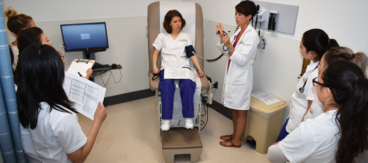 Nursing instructor, right, holds stethoscope in hand while student sits in patient chair with blood pressure cuff on arm