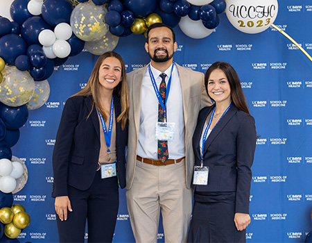 Three medical students – two women and a man – stand next to blue, white and gold balloons and a blue “School of Medicine” backdrop