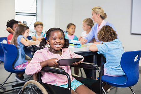 An elementary-aged African American girl in a wheelchair, wearing a pink shirt and blue shorts and holding a tablet, sits in front of a table, around which six students and one teacher sit and talk.