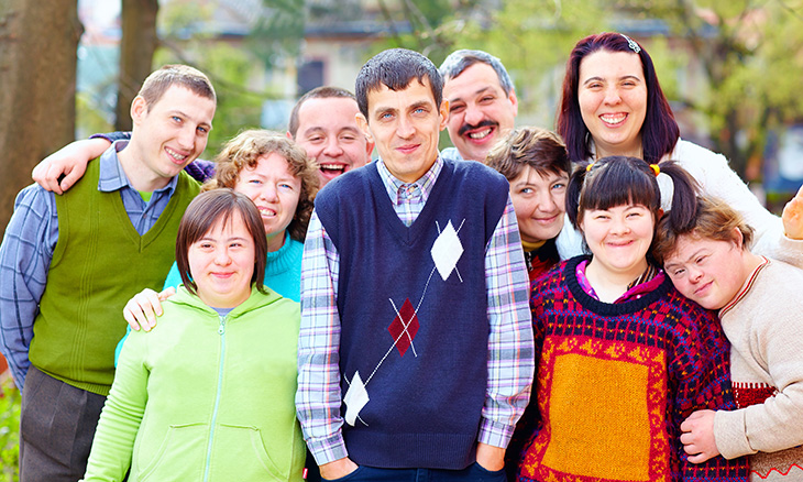 A group of 10 people poses outside for a group photo. Some have Down syndrome or other disabilities. Many have arms around each other. 