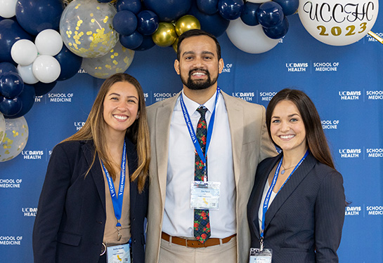 Three medical students – two women and man – stand next to blue, white and gold balloons and a blue “School of Medicine” backdrop