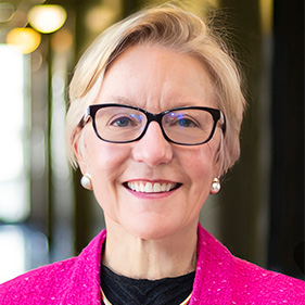 Woman with short blonde hair and horn rimmed glasses wearing pink blazer and black shirt. 
