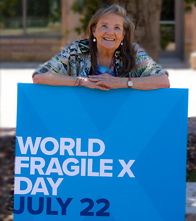 A woman with gray hair stands with her arms resting on a large blue poster that says 