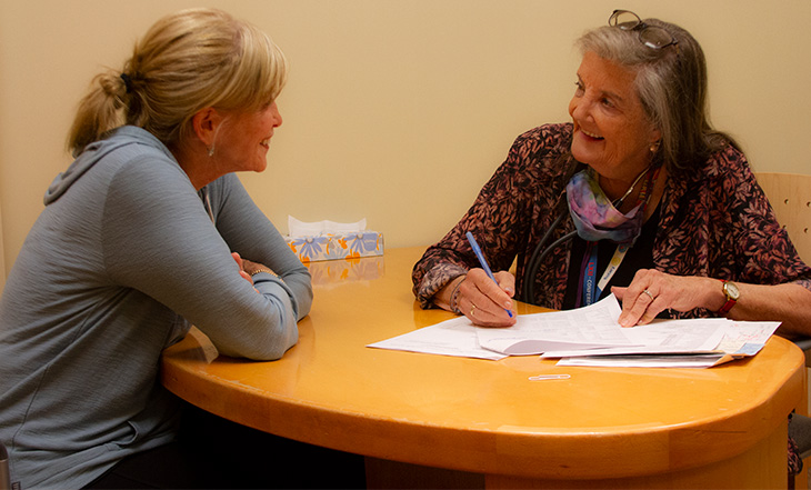 A middle-aged woman with blond hair in a ponytail sits across a table from a woman with gray hair who is taking notes. Assorted papers are on the table between them. 
