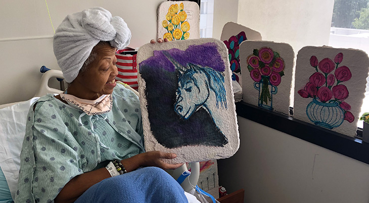 Woman with towel on her head sitting in hospital bed holding a unicorn painting