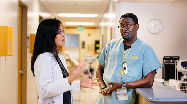 A female doctor with long black hair is wearing a white gown and using her hands to explain something to a medical assistant wearing blue scrubs. The medical assistant has a goatee, is wearing eyeglasses, and is leaning on a counter. There is a round clock on the wall in the background.