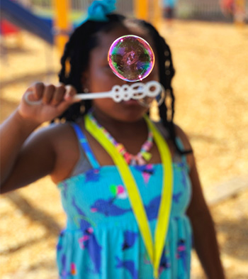 A young girl wearing a blue dress and yellow lanyard blows into a bubble wand as a giant bubble floats on top of the wand, mostly obscuring her face. 