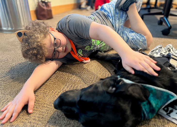 A young boy with brown curly hair, wearing a gray shirt and shorts, lays on the floor next to a black Labrador retriever, petting his fur. 