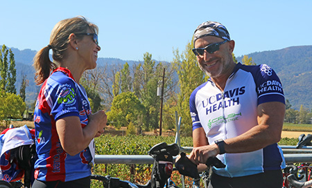 Blonde woman in purple cycling shirt smiling at man wearing bandana, sunglasses and UC Davis Health cycling shirt while leaning on bike.