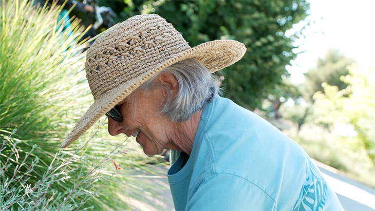 A smiling woman wearing a straw hat, sunglasses and a blue t-shirt is seen leaning down in front of some grassy plants in a garden setting. 