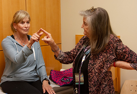 A woman with blond hair in a ponytail, wearing a blue long-sleeved top holds her right arm out and touches fingers with a gray-haired physician