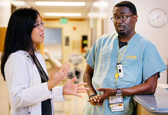 A female doctor with long black hair is wearing a white gown and using her hands to explain something to a medical assistant wearing blue scrubs. 