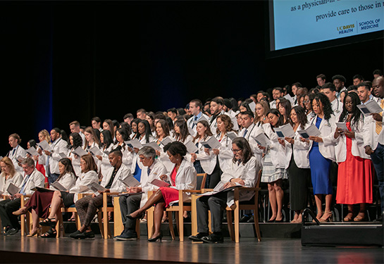 Group of people standing on stage wearing white coats and reading from booklet.