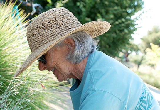 A smiling woman wearing a straw hat, sunglasses and a blue t-shirt is seen leaning down in front of some grassy plants in a garden setting. 