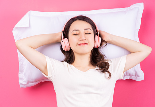 A young Asian woman with her eyes closed and wearing pink headphones is resting on a white pillow against a pink background.