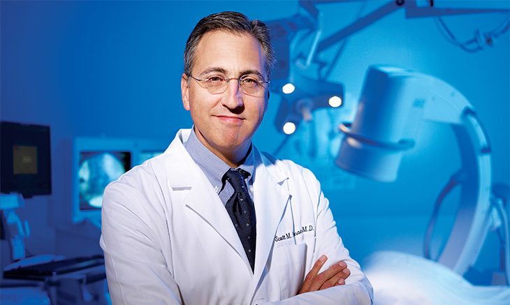 A middle-aged man in glasses and wearing a white physician coat stands with his arms crossed in front of some medical equipment. 