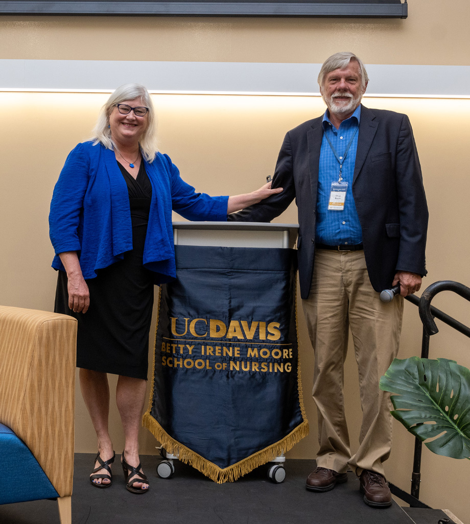 Heather M. Young, left, stands with outstretched arm touching Doug Busch’s arm in front of a podium on stage.